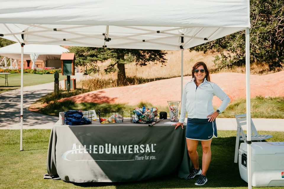 Woman stands at Allied Universal booth on a golf course, smiling. Table displays items under a white tent.