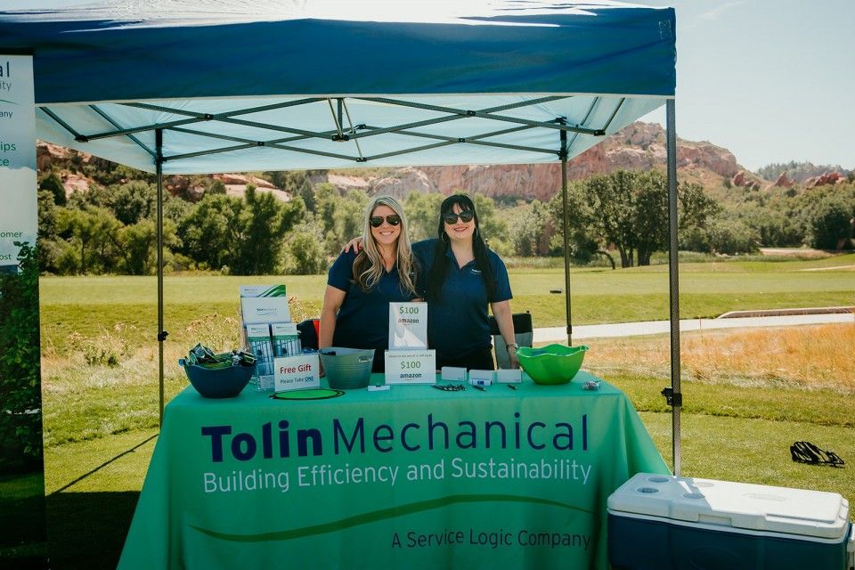 Two women at a Tolkin Mechanical booth on a golf course; green tent, sign, and table cover; sunny day.