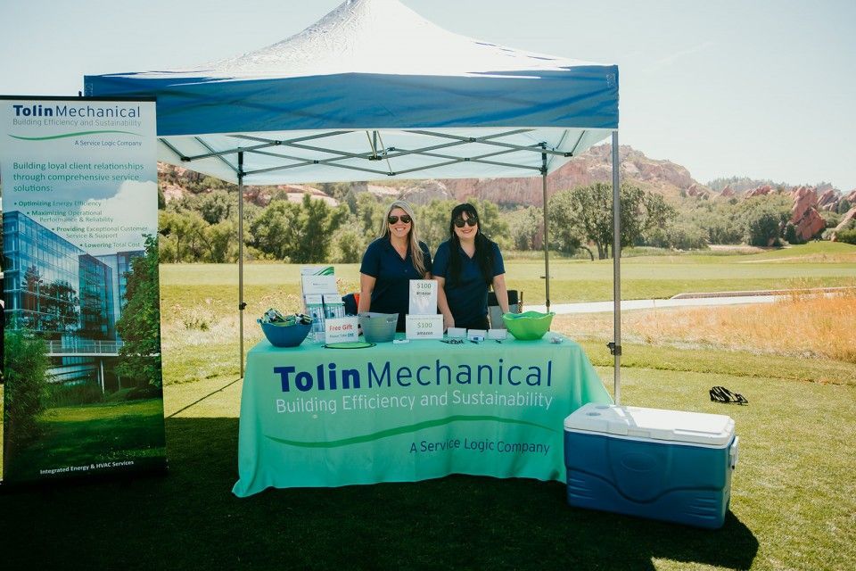 Two women at a Tolon Mechanical booth on a sunny golf course. Green and blue color scheme, food items.