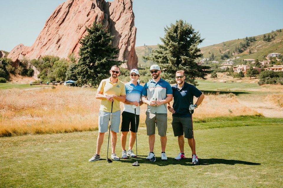 Four men smiling, holding golf clubs on a green course with a large red rock formation in the background.