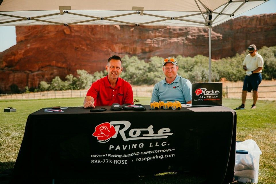 Two men at a Rose Paving LLC booth, table with logo, in outdoor setting, mountain background.