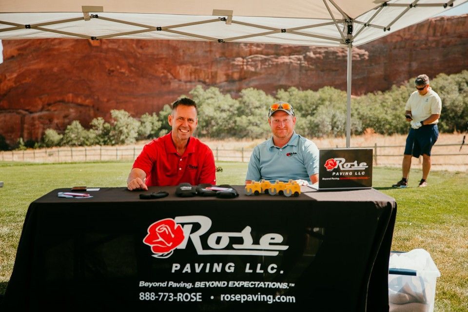 Two men at a Rose Paving LLC booth, red and blue shirts, black tablecloth, red rock backdrop.