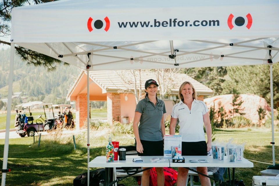 Two women at a BELFOR booth under a tent, smiling. Golf carts and a building are in the background.