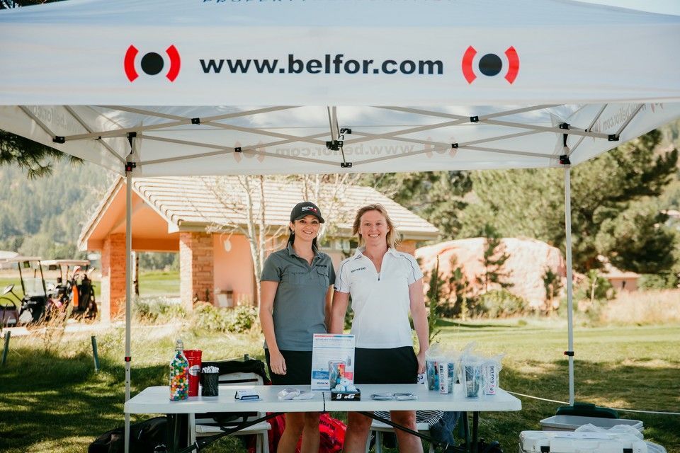 Two women stand behind a table under a BELFOR tent at an outdoor event.
