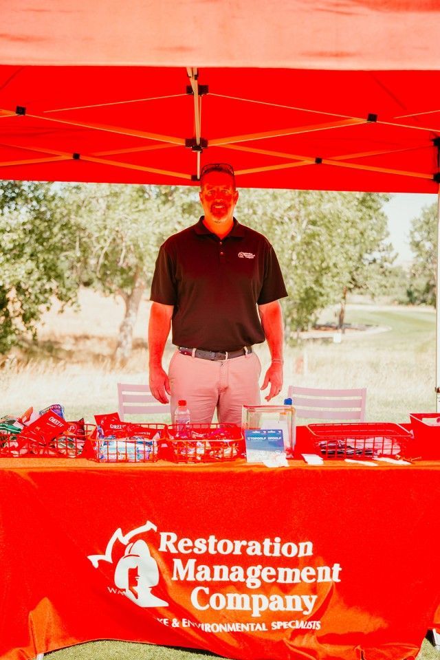 Man standing behind a red table, at a Restoration Management Company booth, outdoors.
