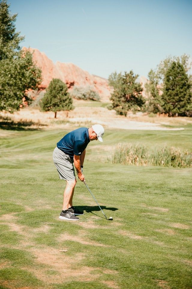 Man swinging a golf club on a green, with red rock formations in the background on a sunny day.
