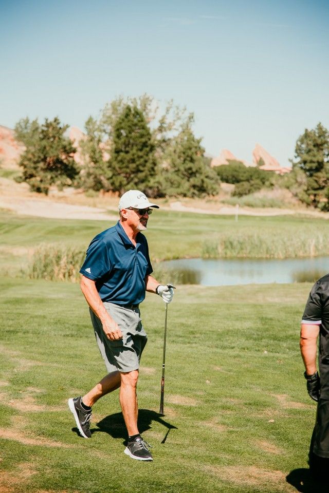 Man in navy polo and shorts walks on a golf course, holding a club. Sunny day.