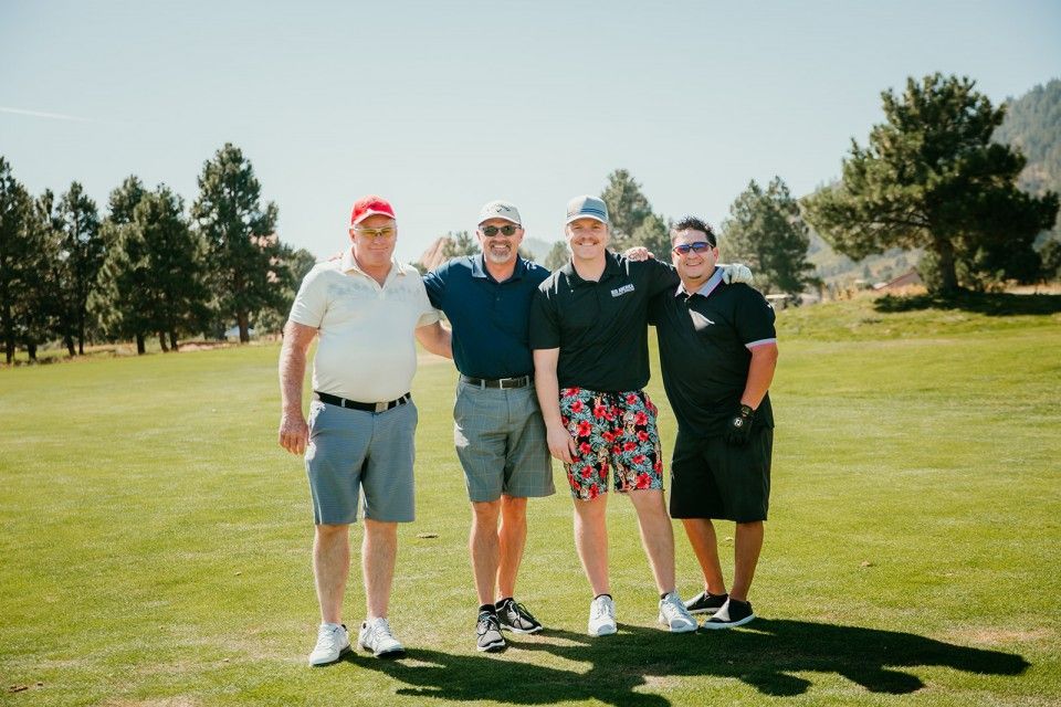Four men standing on a golf course, arms around each other. Bright sunny day.