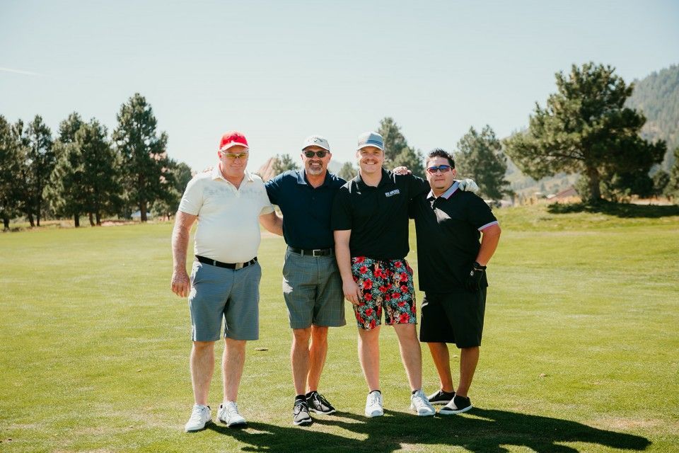 Four men in golf attire pose on a sunny green golf course; trees in the background.