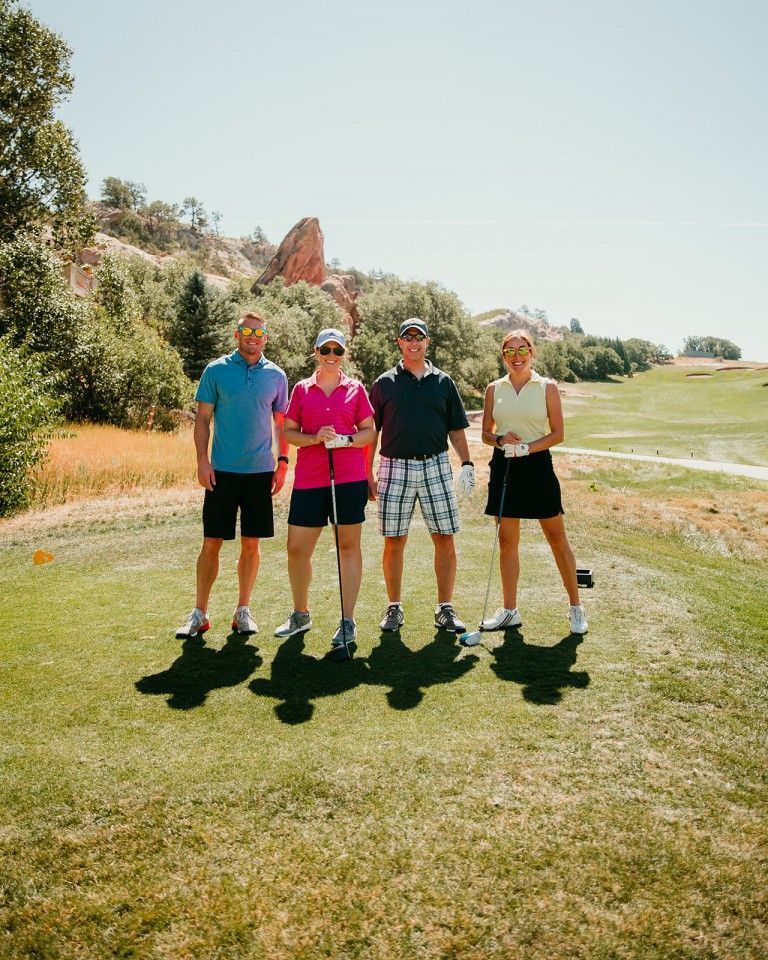 Four golfers on a sunny course: two men and two women, holding golf clubs, with a reddish rock formation in the background.