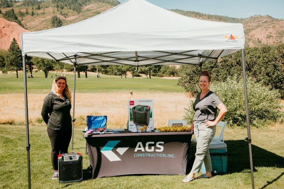 Two women standing at an AGS Construction booth under a white canopy on a golf course, mountains in the background.