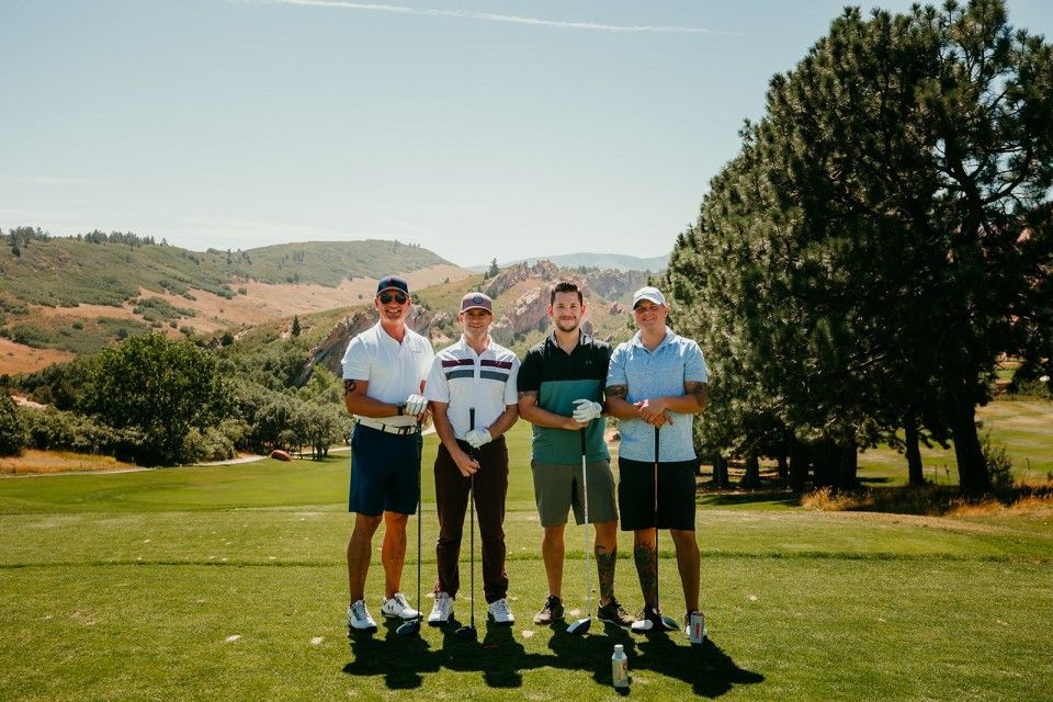 Four men on a golf course, holding clubs. Sunny day, mountains in the background.