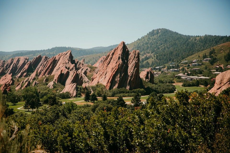 Red rock formations rise above green trees and a small town, under a clear blue sky.