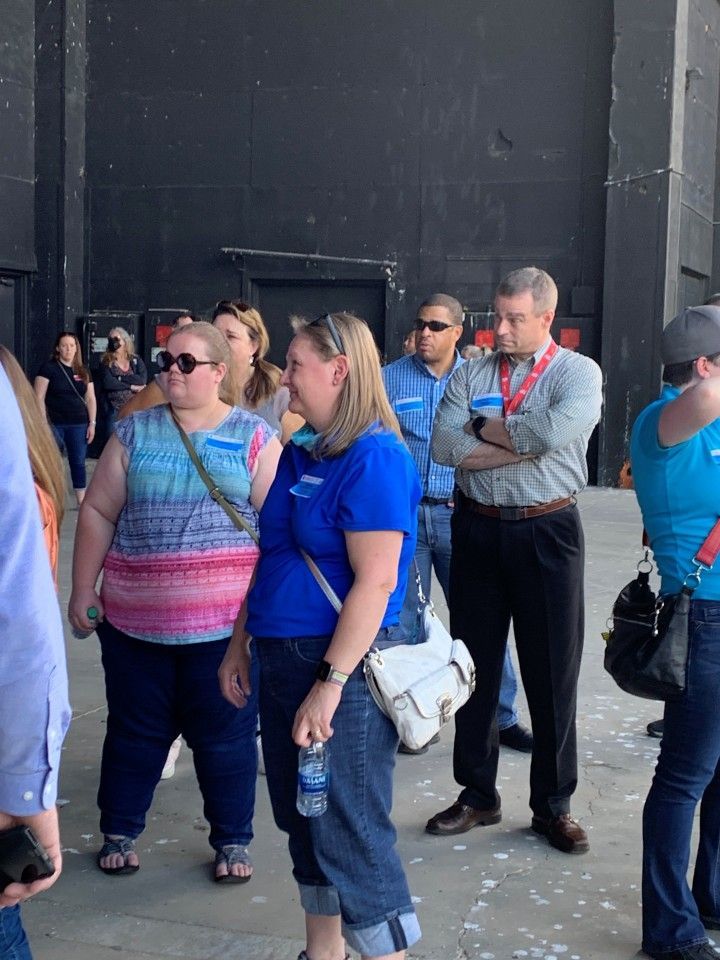Group of people standing outdoors; some are looking at something to their left. The background is a large black wall.