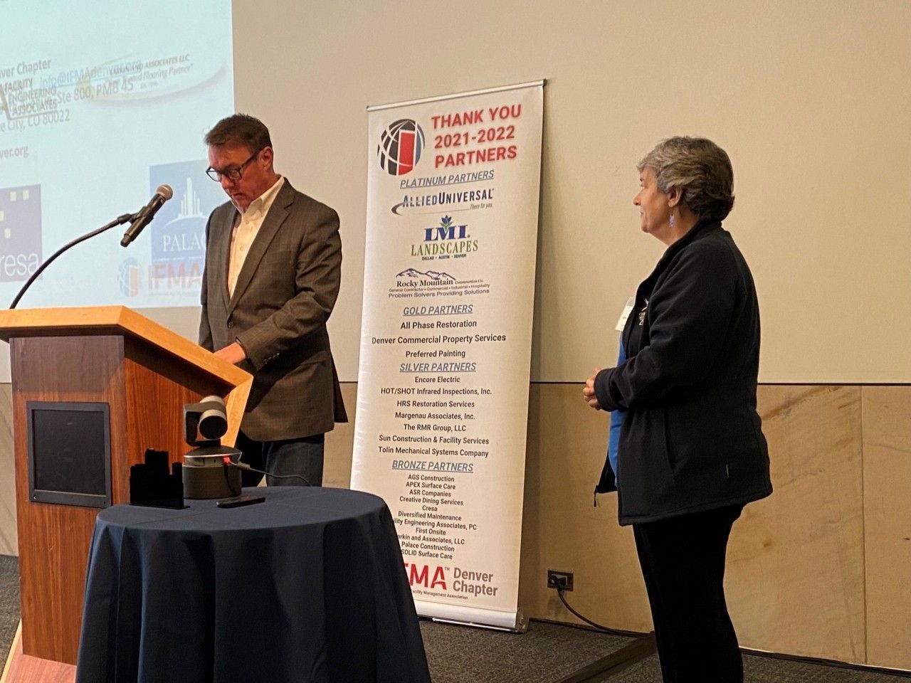 Man speaking at a podium with a woman standing beside a 