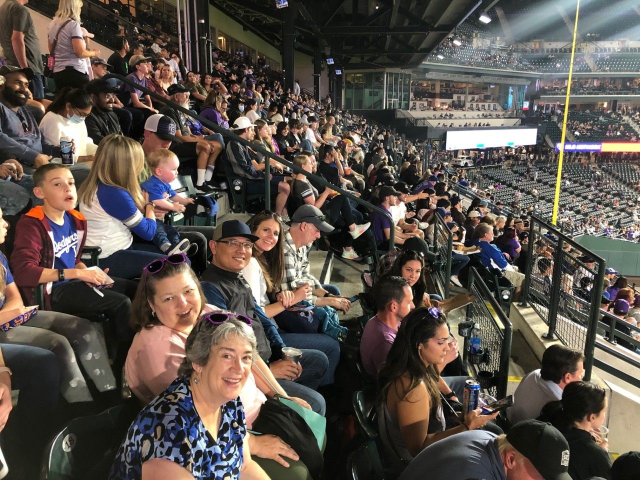 Crowd watches a baseball game from stadium seats. Some smile, wave. Mostly dark setting with overhead lights.