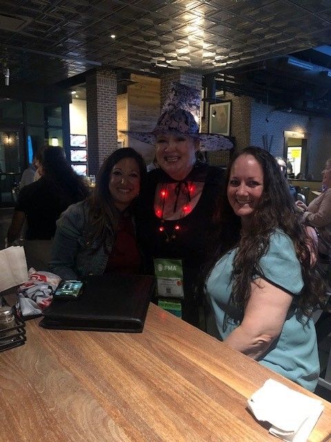 Three women at a restaurant, one in witch costume. They smile at the camera, posing near a wooden table.
