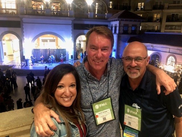 Three people smiling for a photo outdoors. A man in the middle has his arms around the others. Stage in background.