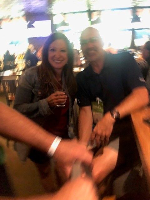 A woman and a man smiling at the camera, holding a drink, at a bar. Arms clink hands in foreground.