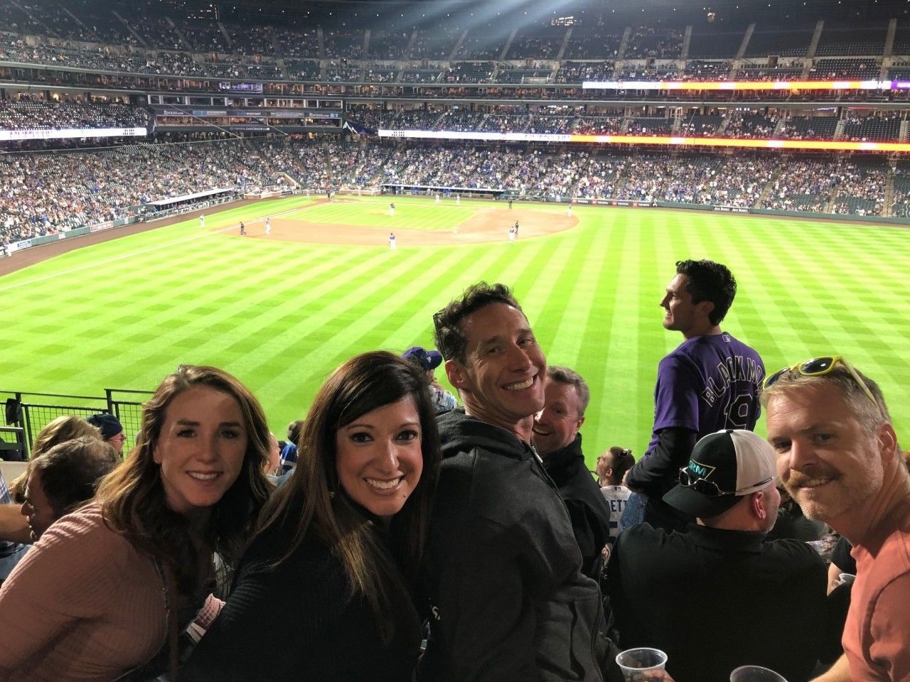 People smile at a baseball game, with the field and packed stadium in the background.