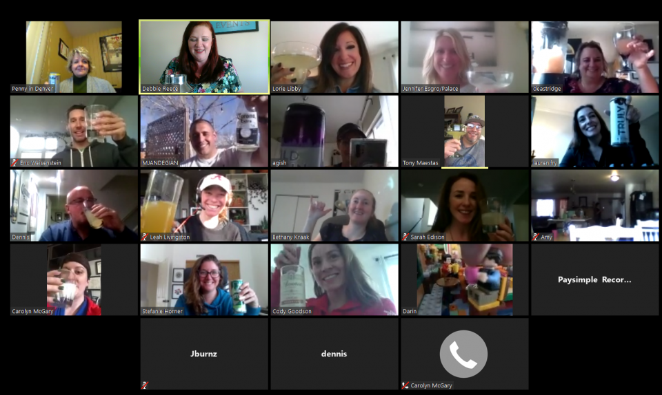 Group of people on a video call, raising drinks and smiling, indoors.
