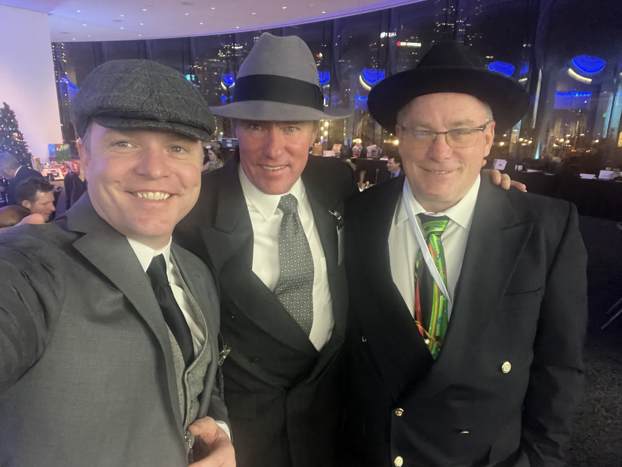 Three men in suits and hats posing indoors. One wears a grey cap, a second a grey fedora, and the third a black hat.