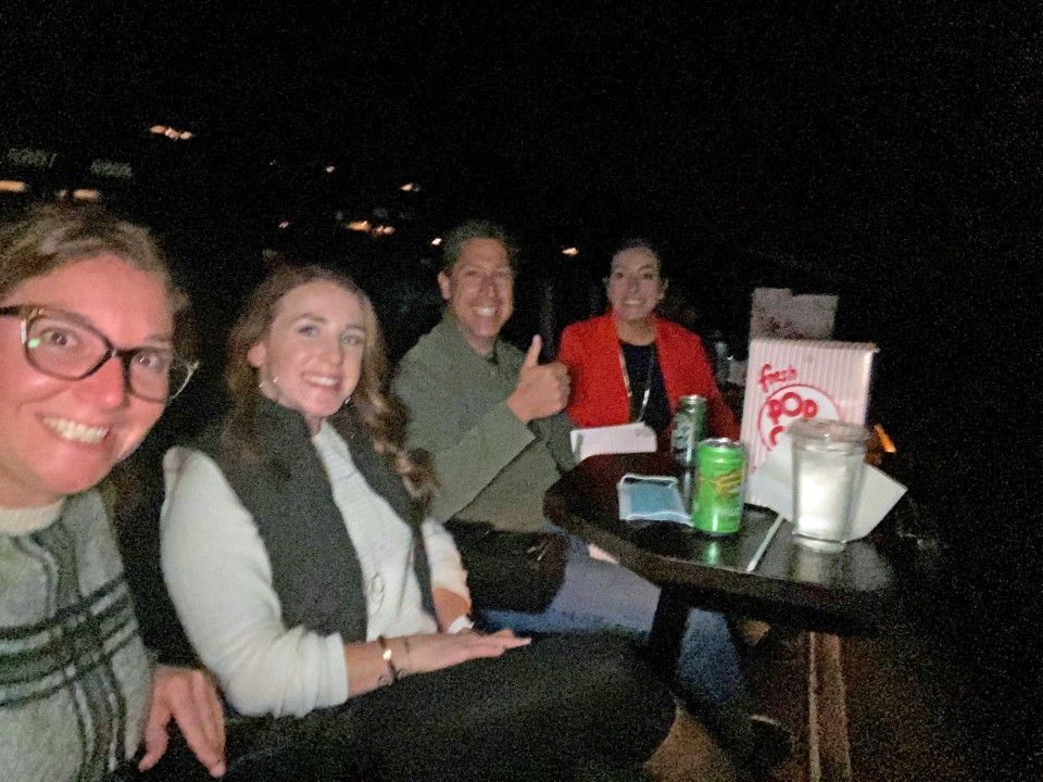 Four people seated at a small table, smiling.  One gives a thumbs-up.  Dark room, with a soda and a box on the table.