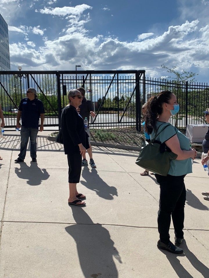 People standing outside a gated area on a sunny day. Some are wearing masks and carrying bags.