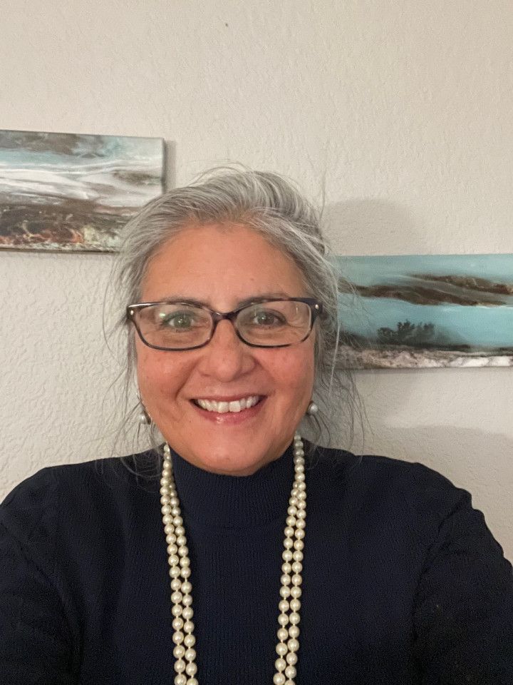 Woman with gray hair and glasses smiles, wearing a pearl necklace and dark sweater. Background has landscape paintings.