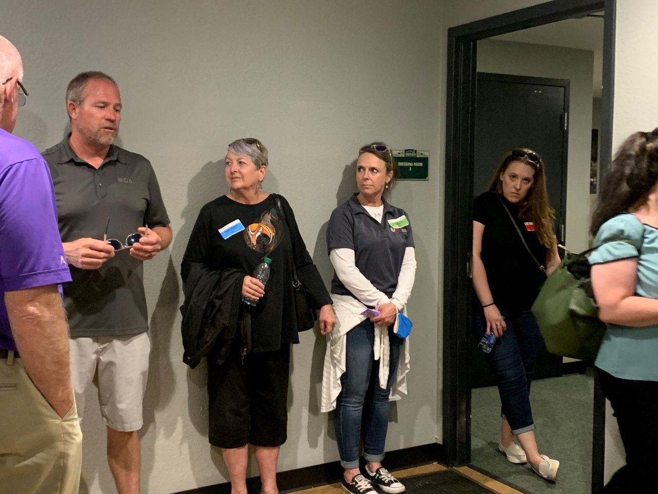 People standing in a hallway. Some are talking, others are looking forward. Dark door and a green sign are visible.