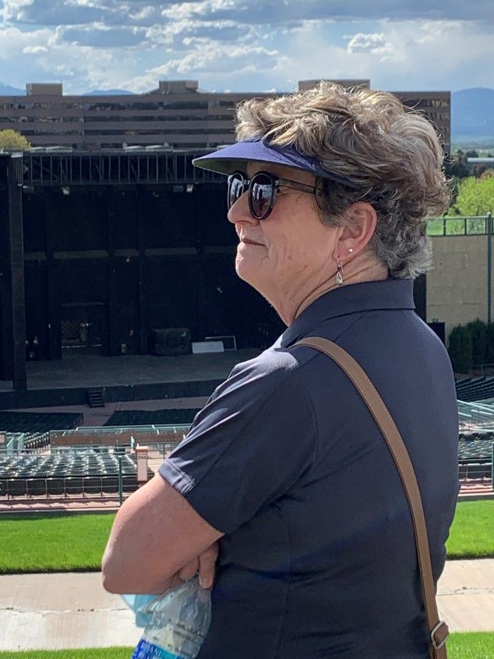 Woman wearing sunglasses and visor, looking at an outdoor stage; arms crossed.