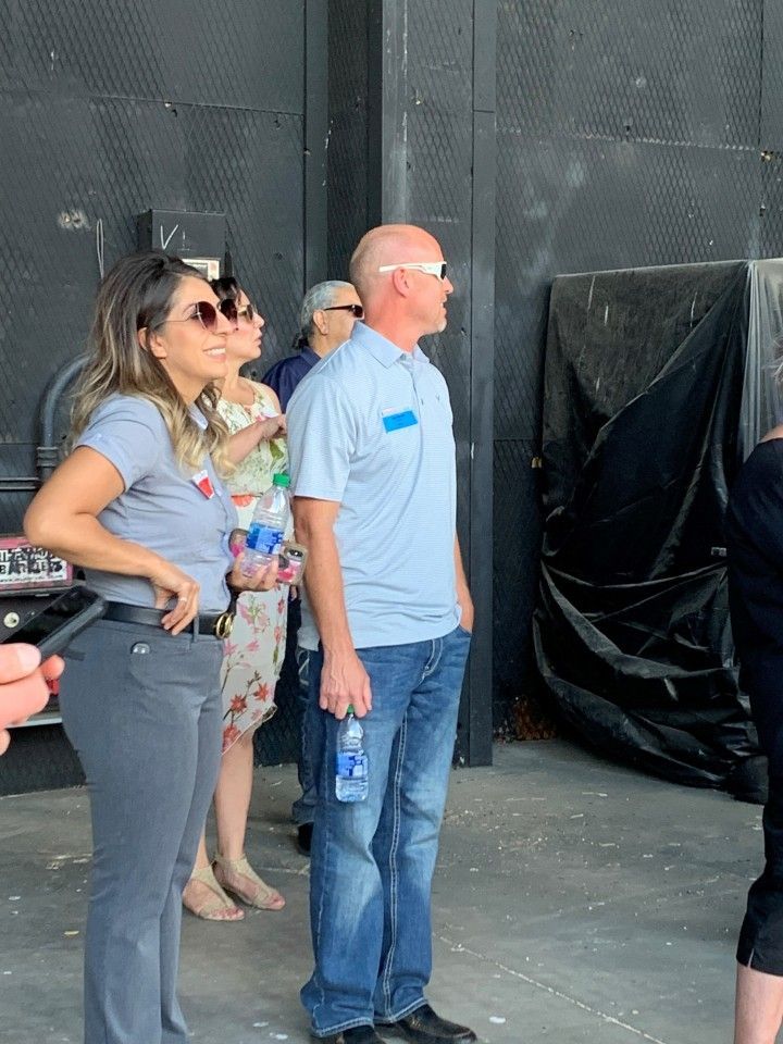 People, some in sunglasses, stand near a dark structure, looking right. Man in blue shirt holds a water bottle.