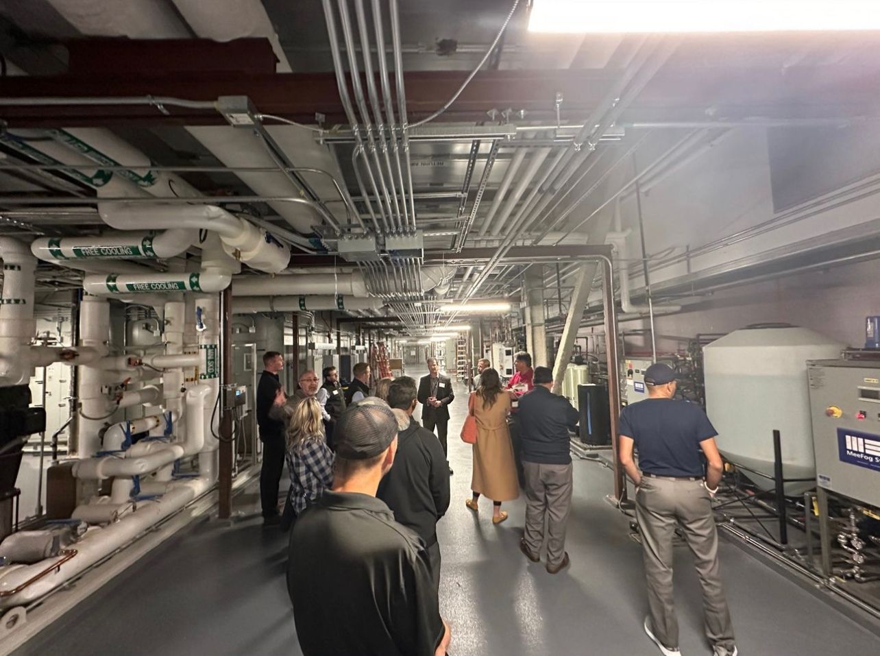 People tour an industrial-looking room with pipes and equipment. They are walking and looking around.