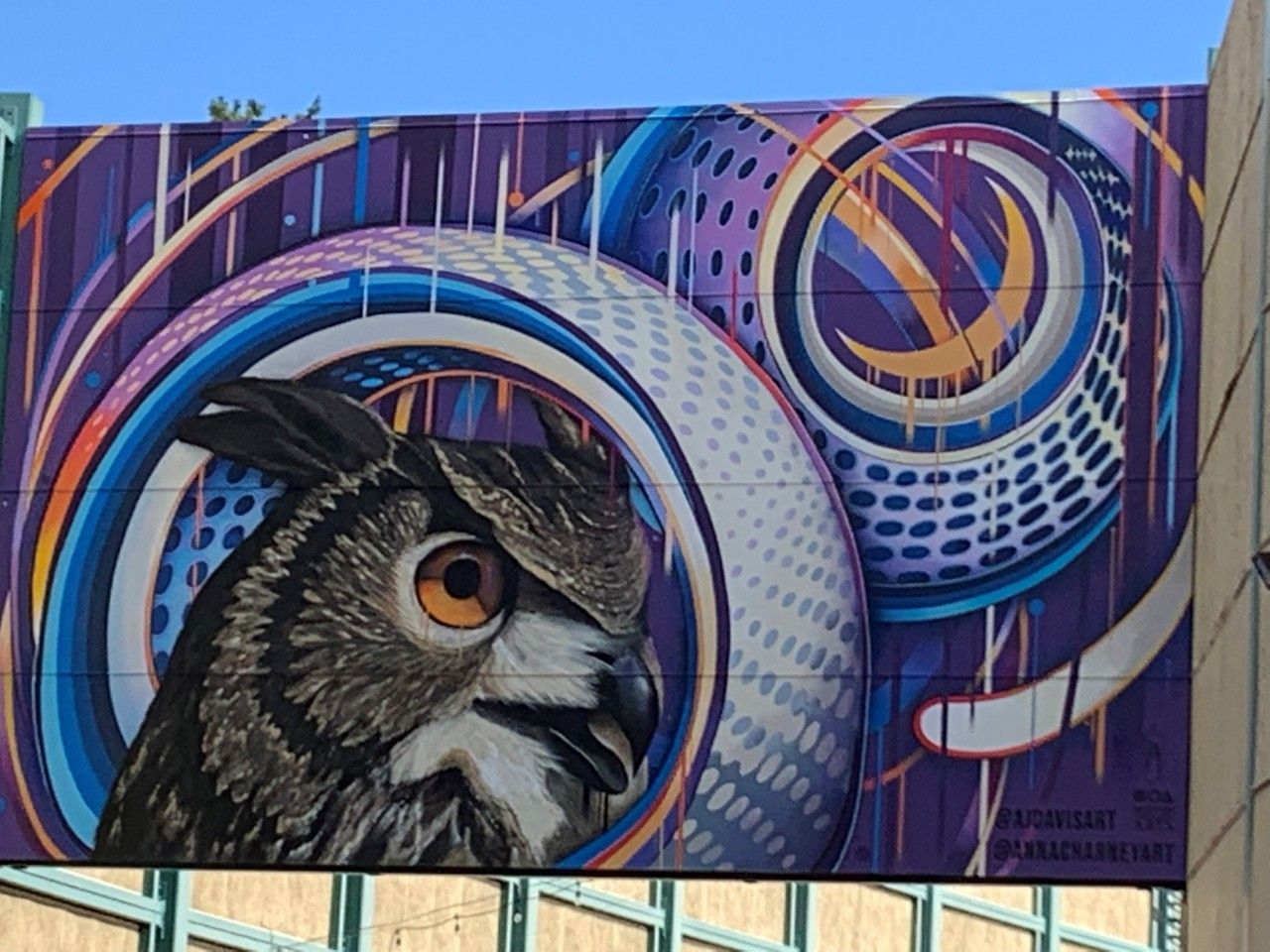 Mural of an owl with orange eyes, surrounded by colorful swirls and circles, on a purple and blue background.
