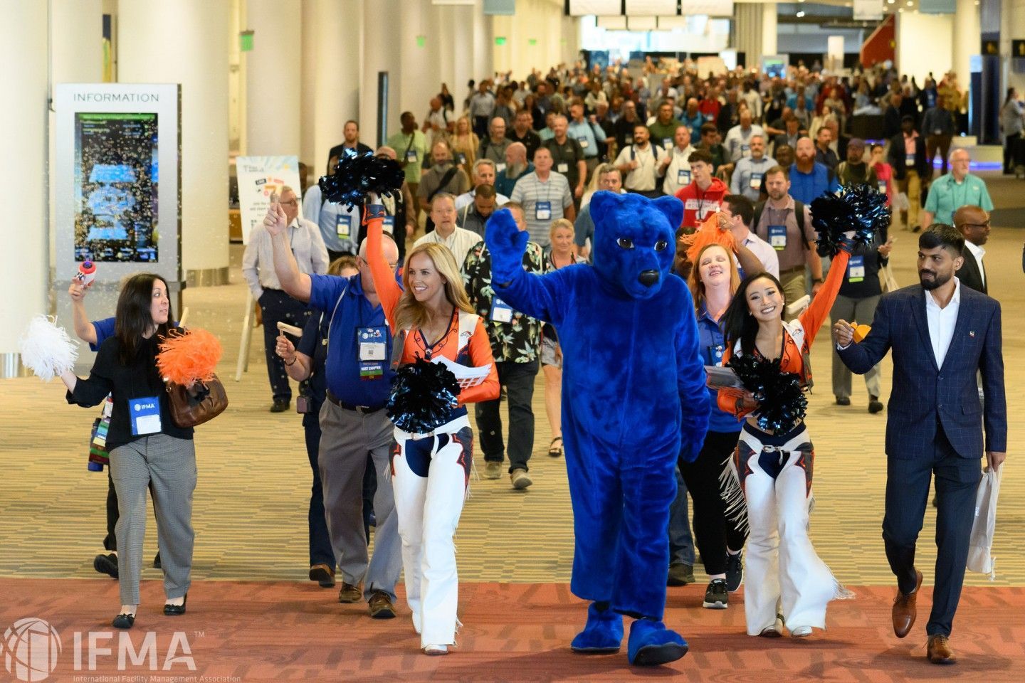 Mascot and cheerleaders lead a crowd through a convention center hallway.