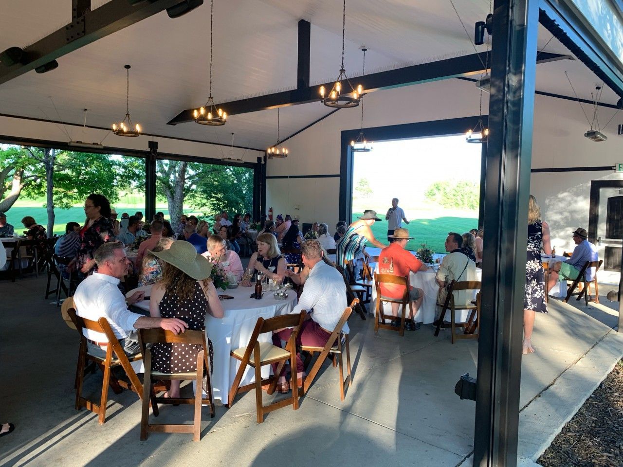 Guests seated at tables under an open-air pavilion. People eat, converse, and enjoy a gathering.
