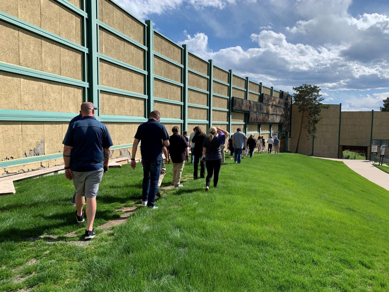 A group walks along a grassy area next to a large noise barrier on a sunny day.