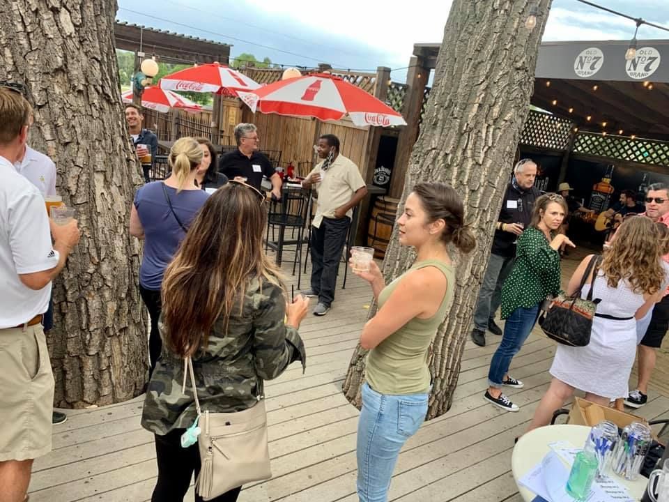 People socializing outdoors on a wooden deck, trees and red umbrellas visible.