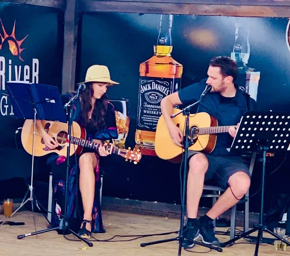 Two musicians performing with acoustic guitars on a stage; woman in a hat, man singing, Jack Daniel's backdrop.