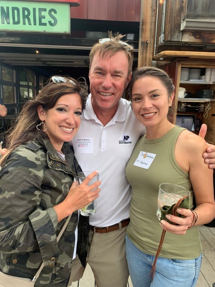 Three people smile, posing for a photo outdoors. Man in polo shirt flanked by two women, all holding drinks.