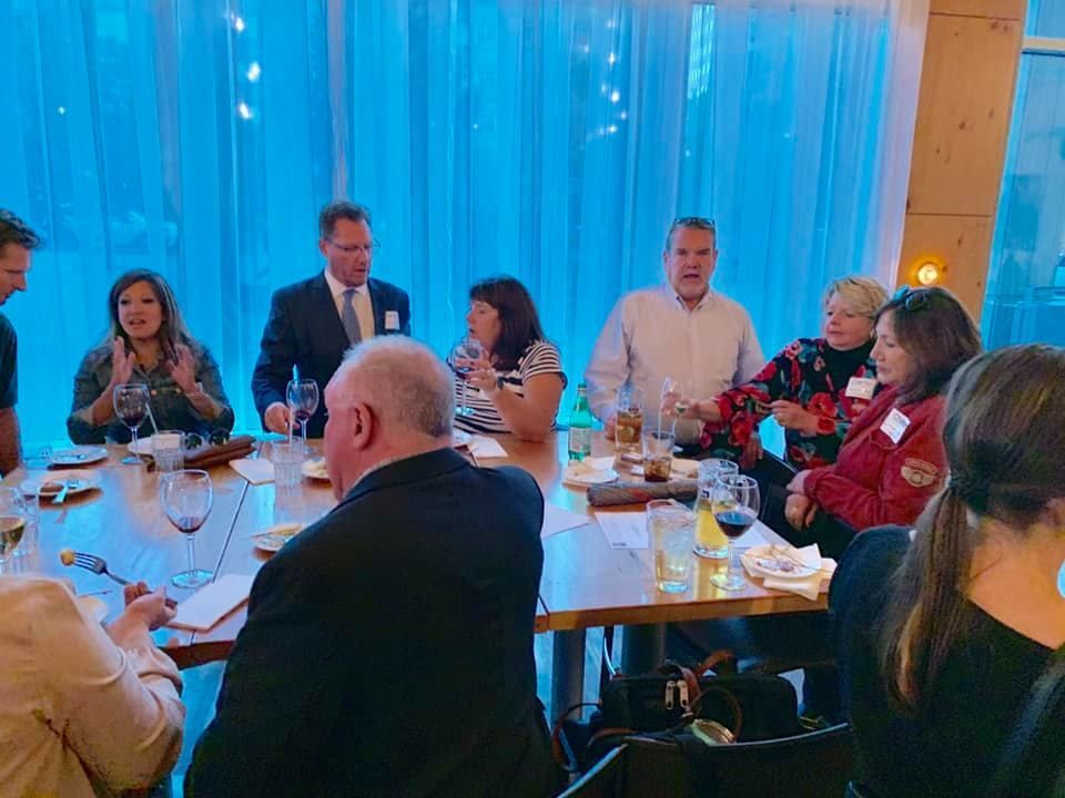 People seated at a long table, dining. A window is in the background, with blue lighting.