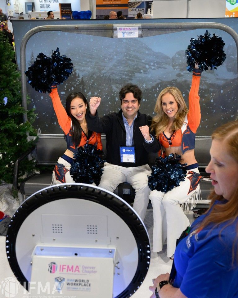 Man poses with two cheerleaders, holding pom-poms, in front of a photo booth, at a conference.