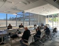 People seated at a conference table in a modern room with large windows; a presentation is in progress.
