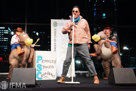 Man singing on stage, flanked by two people in donkey costumes. City backdrop.
