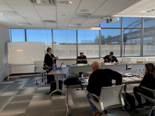 A woman presents at a table with other people in a meeting room with large windows.