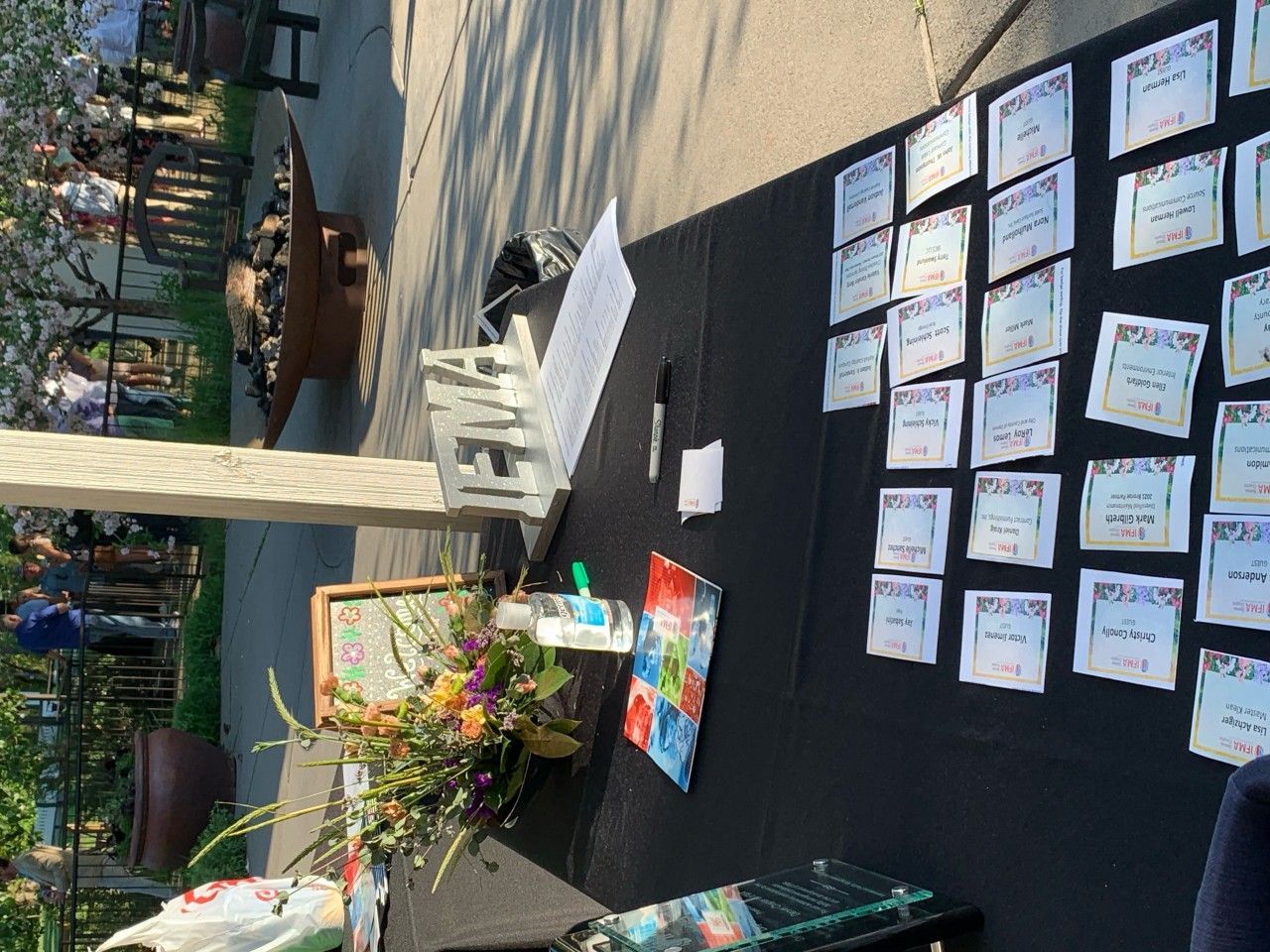 Table with name cards, flowers, and a sign. Guests in the background. Outdoor setting.