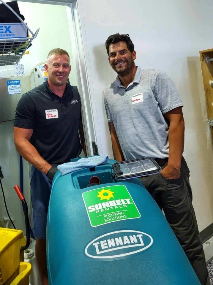Two men pose with a teal Tennant floor scrubber. One wears a black shirt, the other a gray polo. Bright interior.