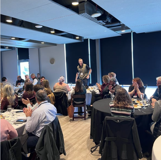 People applauding a man speaking at a business event in a room with round tables and dark window shades.