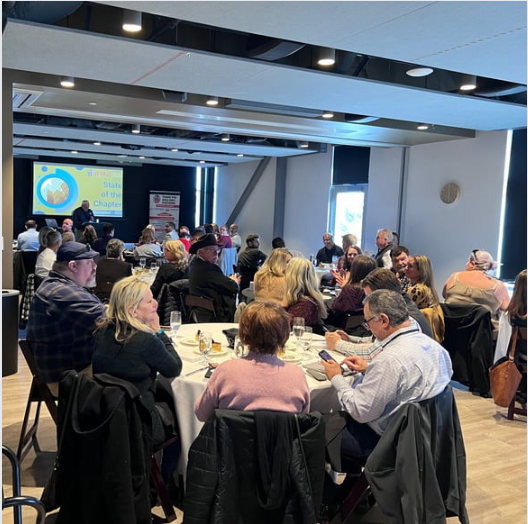 People seated at round tables in a brightly lit room with a presentation screen.