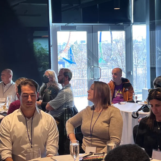 People seated at tables in a conference room. Some look at a presentation. Daylight through the windows.